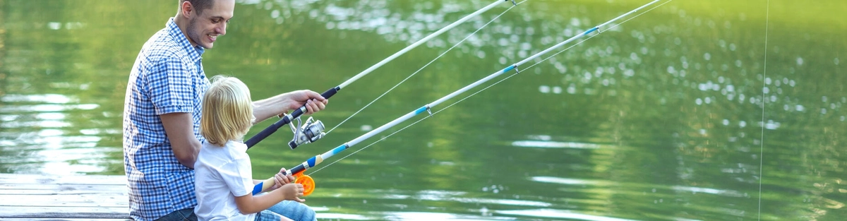 A man and a young child sit on a wooden dock, smiling and fishing together with rods, facing a calm green lake surrounded by nature on a sunny day.