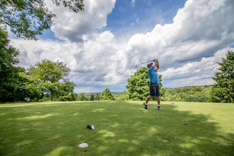 A person in a blue shirt swings a golf club on a lush, green golf course under a partly cloudy sky, surrounded by trees and distant hills.