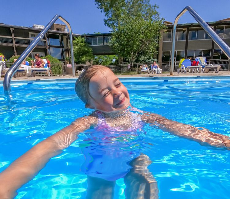 A young child smiles and swims in a bright blue outdoor pool on a sunny day. People relax on lounge chairs and a building with balconies is visible in the background.