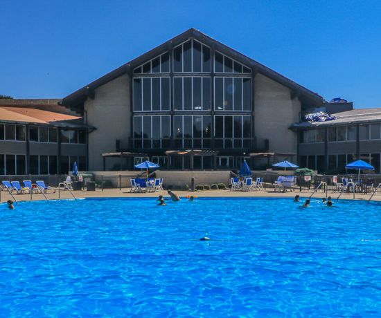 A large outdoor swimming pool with people swimming, surrounded by lounge chairs and blue umbrellas, sits in front of a multi-story lodge-style building with many windows under a clear blue sky.