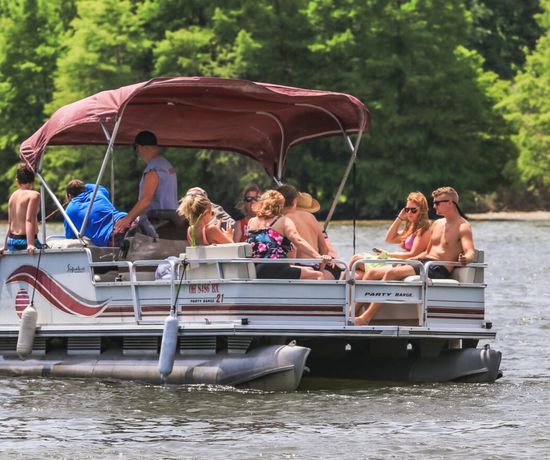 A group of people relax and socialize on a pontoon boat with a maroon canopy, floating on a lake surrounded by lush green trees. The weather is sunny, and most people are wearing swimsuits and sunglasses.