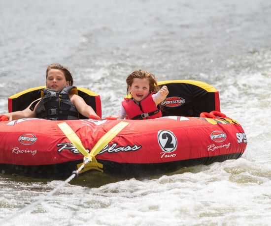 Two children wearing life jackets ride a red inflatable tube being pulled across the water. The child on the right is smiling and raising a fist, while the other sits calmly. Water splashes behind them.