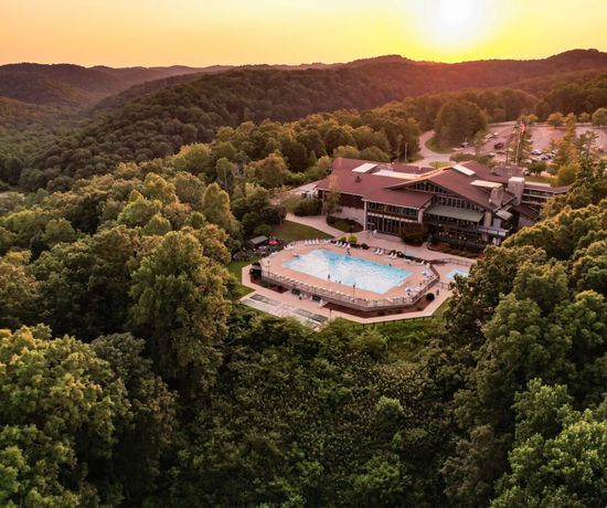 Aerial view of a large lodge with an outdoor swimming pool surrounded by lush green forested hills at sunset, with the sun low on the horizon.