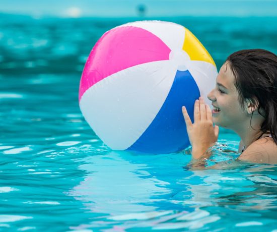 A smiling girl in a swimming pool holds a colorful beach ball. The water is bright blue, and she appears happy and playful as she looks at the ball.