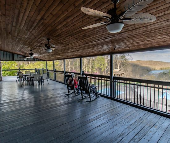 A spacious covered wooden porch with ceiling fans, rocking chairs, and a dining table overlooks a pool, trees, and a river with hills in the distance under a partly cloudy sky.