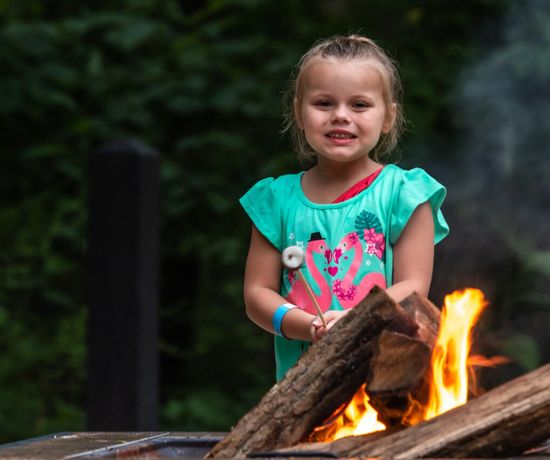 A young girl in a turquoise shirt holds a marshmallow on a stick over a campfire, smiling, with green trees in the background.