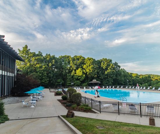 A large outdoor swimming pool with clear blue water, surrounded by lounge chairs and umbrellas. A few people enjoy the pool area, with trees and greenery in the background under a partly cloudy sky.