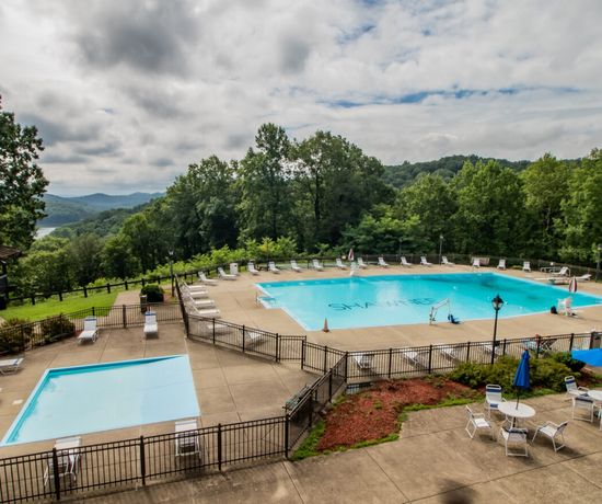 A large outdoor pool and a smaller wading pool are surrounded by lounge chairs and tables with umbrellas, set on a concrete deck amidst lush green trees and distant hills under a partly cloudy sky.